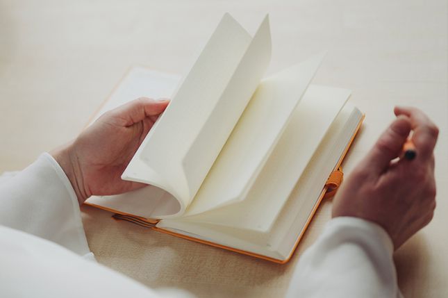 Eine Frau blättert in einem Buch mit leeren Seiten und hält einen orangen Stift in der Hand.